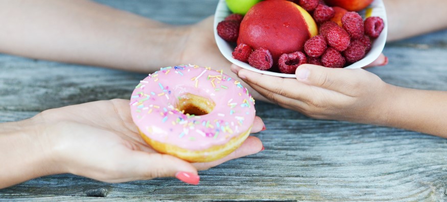 adult holding doughnut in one had and passing a bowl of fruit to child with other hand