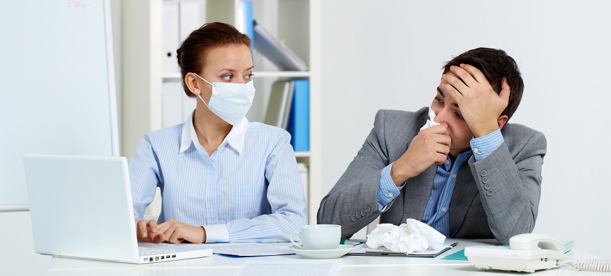 woman wearing mask sat at a desk next to a man blowing nose